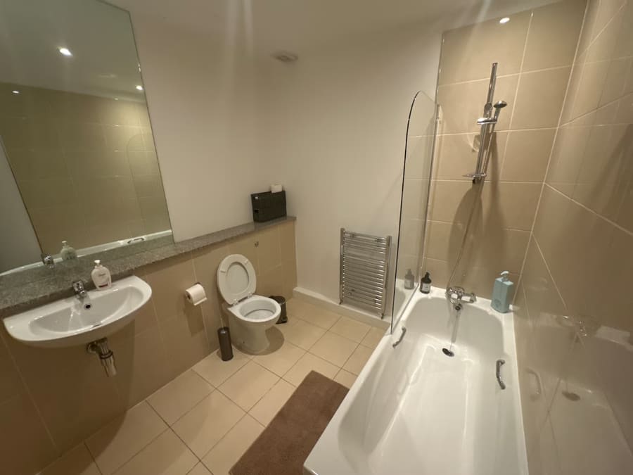 A dated bathroom before redesign, with beige wall tiles, a pedestal basin and a bath shower screen